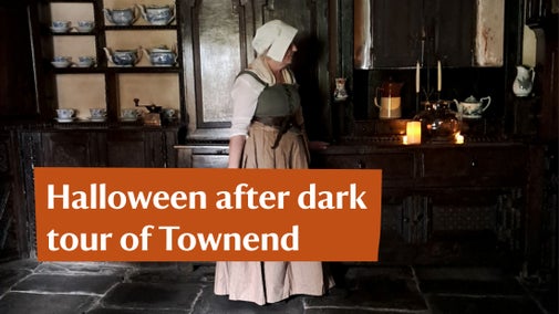 A woman in period clothing standing in a dark kitchen lit by candlelight
